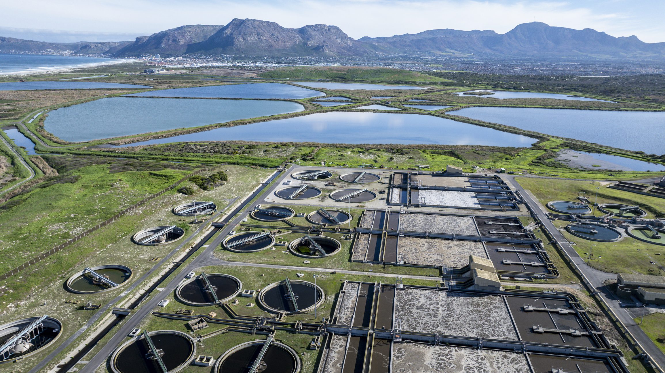 Wide aerial view of an industrial waste water treatment plant with circular clarifier tanks, stabilization ponds and maturation ponds beyond.